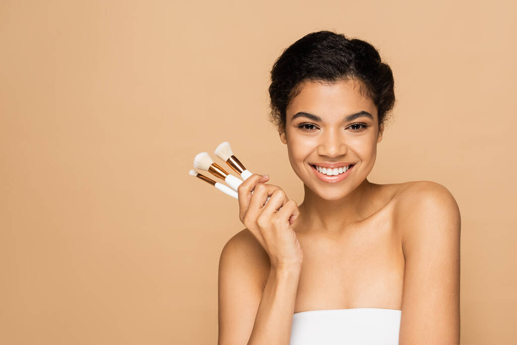 happy young african american woman with bare shoulders holding cosmetic brushes isolated on beige  - Photo, Image