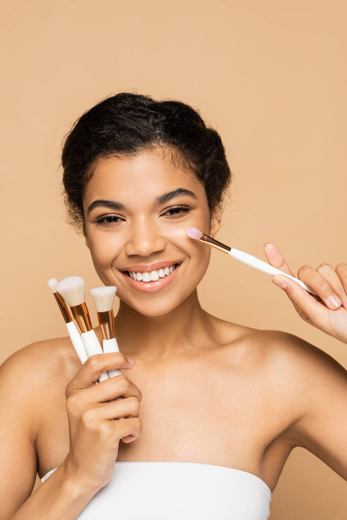 brunette african american woman with bare shoulders applying face powder and holding cosmetic brushes isolated on beige  - Photo, Image