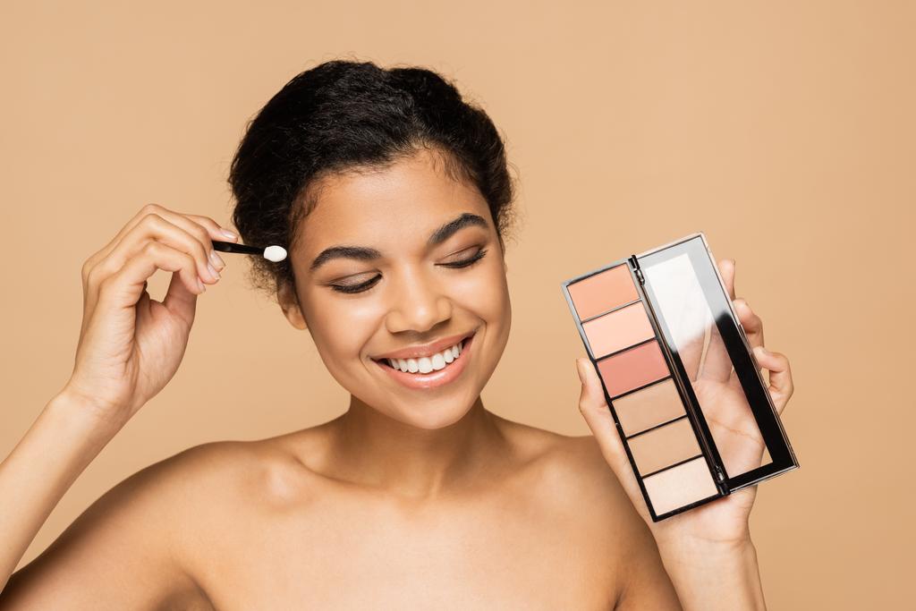 szczęśliwa Afroamerykanka z gołymi ramionami trzymając pędzel kosmetyczny i rumieniec palety izolowane na beżu  - Zdjęcie, obraz