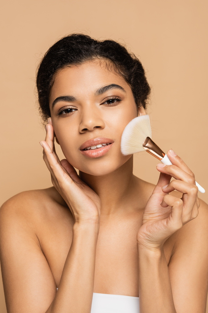 mujer americana bastante africana con hombros desnudos aplicando polvo facial con cepillo cosmético aislado en beige  - Foto, Imagen