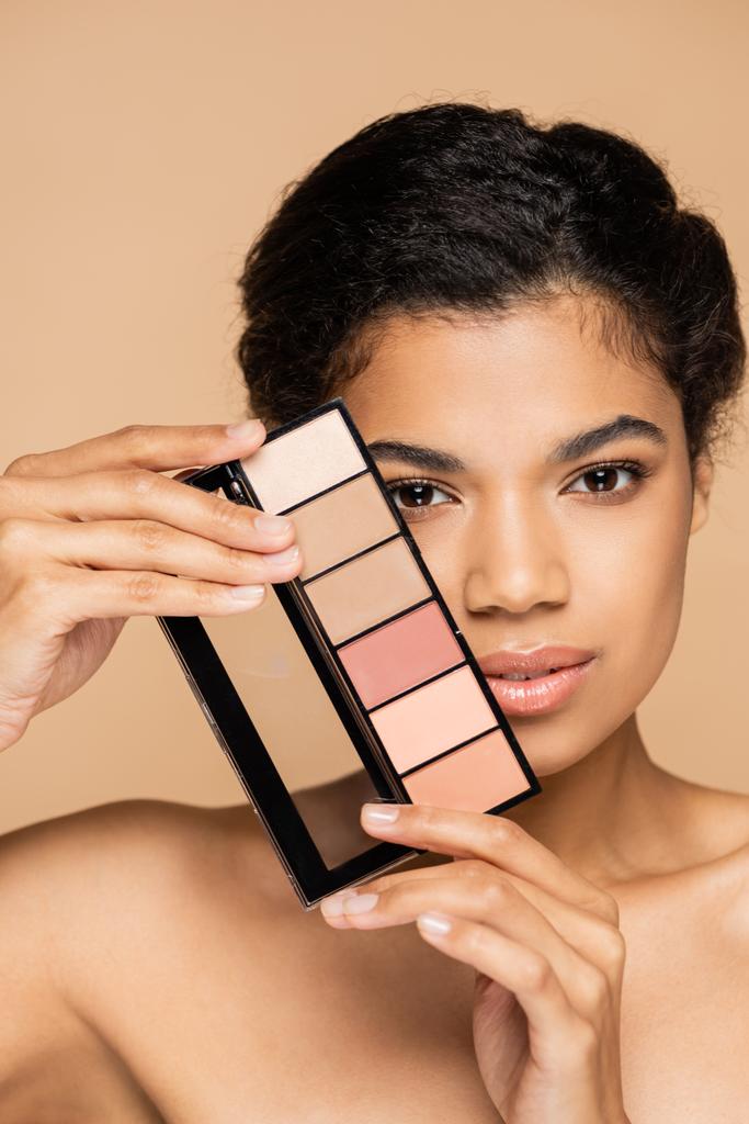 giovane donna afroamericana con spalle nude tenendo tavolozza cosmetica isolata sul beige  - Foto, immagini