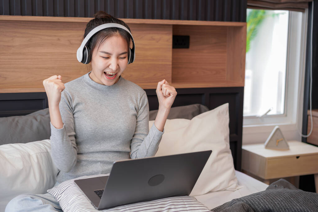 Молода азіатка з навушниками піднімає руки після отримання електронної пошти хороші новини, сидячи на роботі і слухаючи музику з ноутбука на ліжку в домашній спальні
 - Фото, зображення