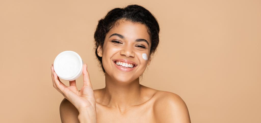 smiling african american woman with bare shoulders holding container with cosmetic cream isolated on beige, banner - Photo, Image