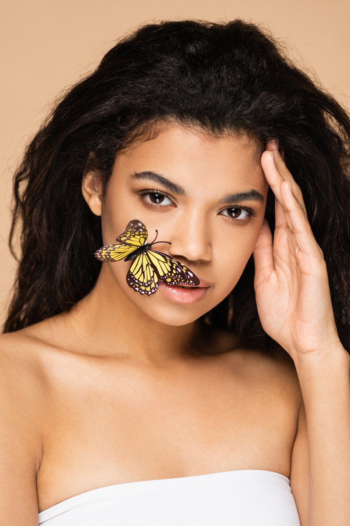 jong Afrikaans amerikaans vrouw met vlinder op gezicht kijken naar camera geïsoleerd op beige - Foto, afbeelding