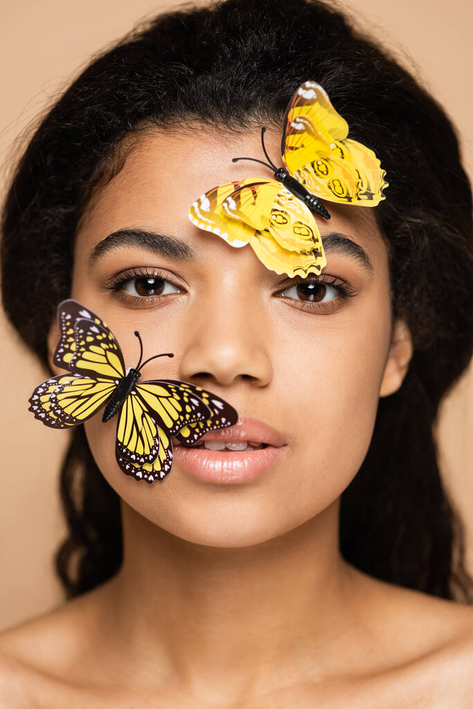 молода афроамериканська жінка з декоративними метеликами на обличчі дивиться на фотоапарат, ізольований на бежевих. - Фото, зображення