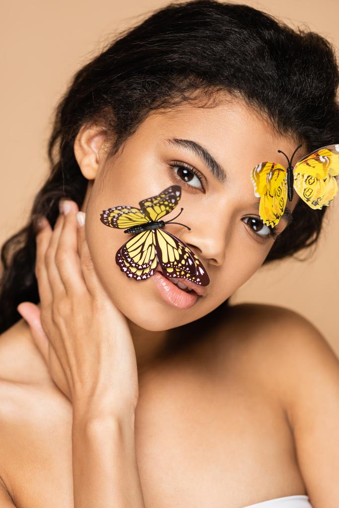 afro-américaine jeune femme avec des papillons décoratifs sur le visage en regardant la caméra isolée sur beige - Photo, image