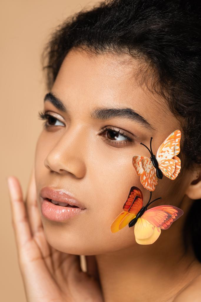 крупным планом брюнетки африканской американки с декоративными бабочками на щеке, изолированными от бежевого - Фото, изображение