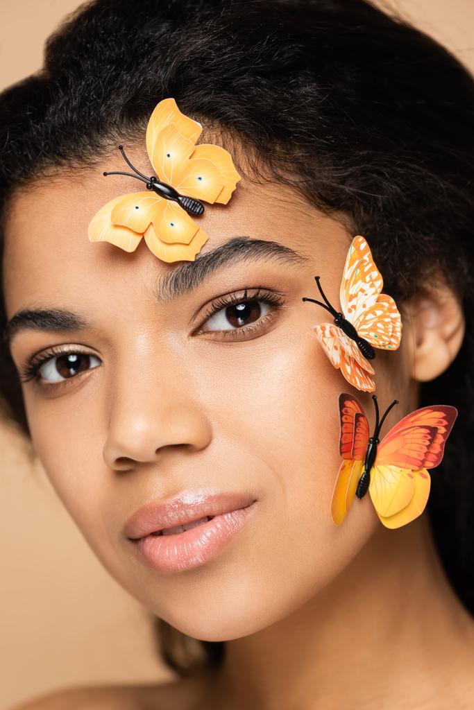 κοντινό πλάνο της νεαρής Αφροαμερικανής γυναίκας με πεταλούδες στο πρόσωπο κοιτάζοντας κάμερα απομονωμένη σε μπεζ - Φωτογραφία, εικόνα