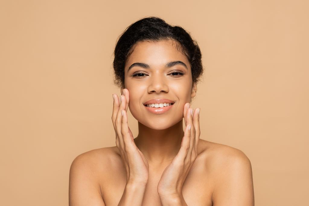 happy african american woman with bare shoulders touching clean face isolated on beige  - Photo, Image