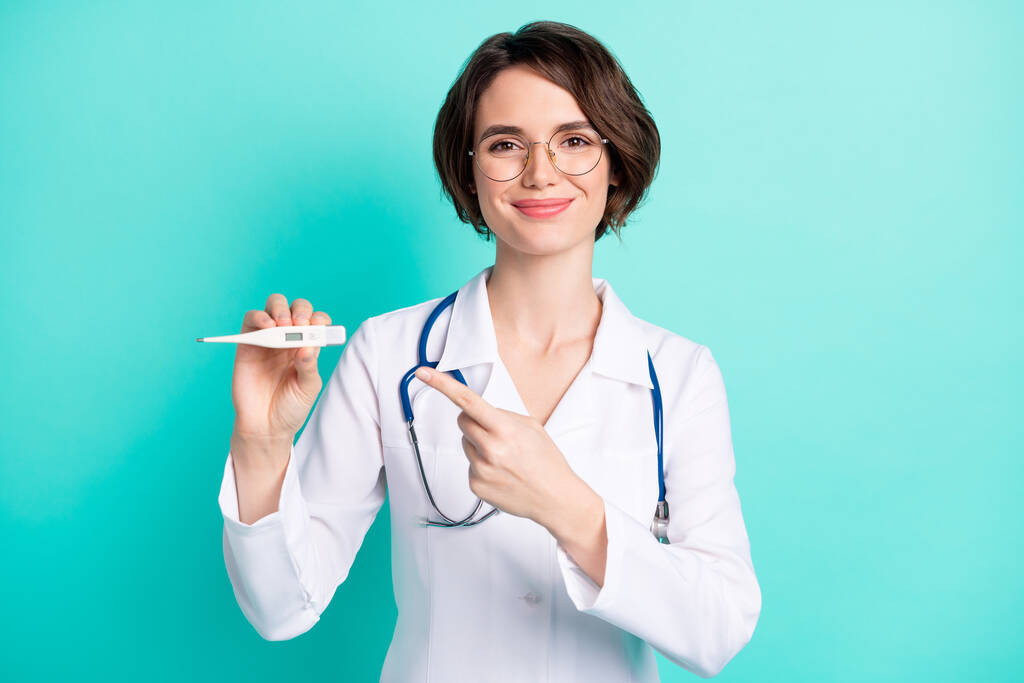 Фотопортрет веселой женщины-врача в белом пальто, указывая пальцем термометр изолирован яркий бирюзовый цвет фона - Фото, изображение