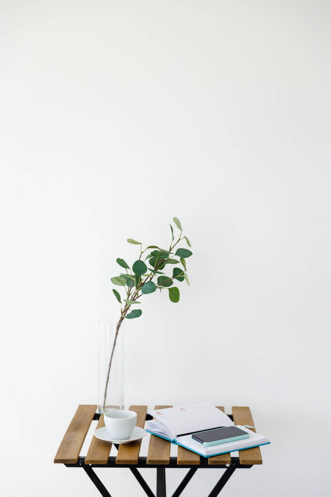 Дерев'яний стіл на фоні білої стіни. Ваза з евкаліптовою гілкою, білою чашкою та смартфоном з блокнотом. Робоче місце фрілансера
 - Фото, зображення