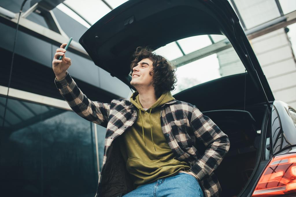 Vista de bajo ángulo del hombre tomando selfie en el teléfono inteligente cerca del maletero del coche al aire libre  - Foto, imagen