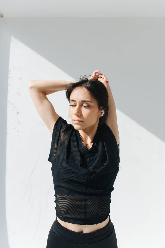 mujer armenia soñadora de pie con las manos detrás de la cabeza cerca de la pared blanca con sombras - Foto, Imagen