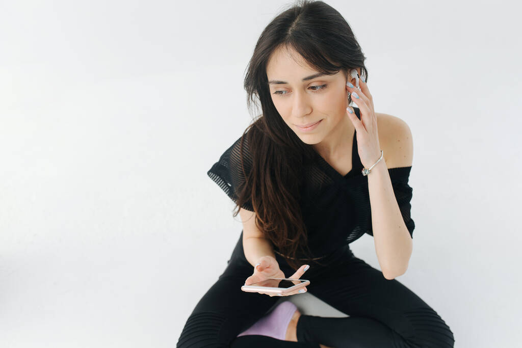 arménská žena v černém oblečení upravující sluchátka při sezení s mobilním telefonem na bílém - Fotografie, Obrázek
