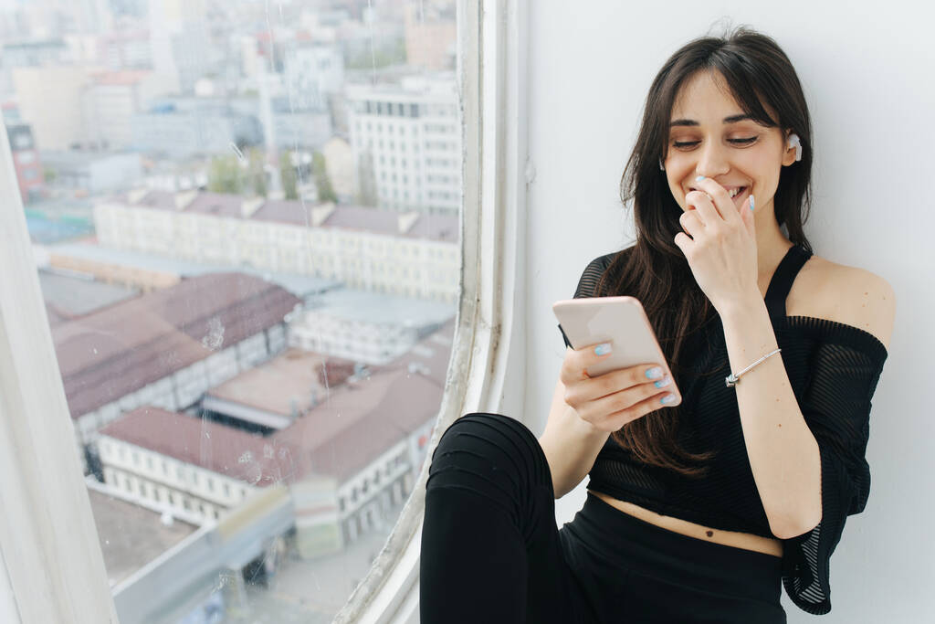 glückliche armenische Frau, die Mund mit der Hand bedeckt, während sie Smartphone in Fensternähe benutzt - Foto, Bild