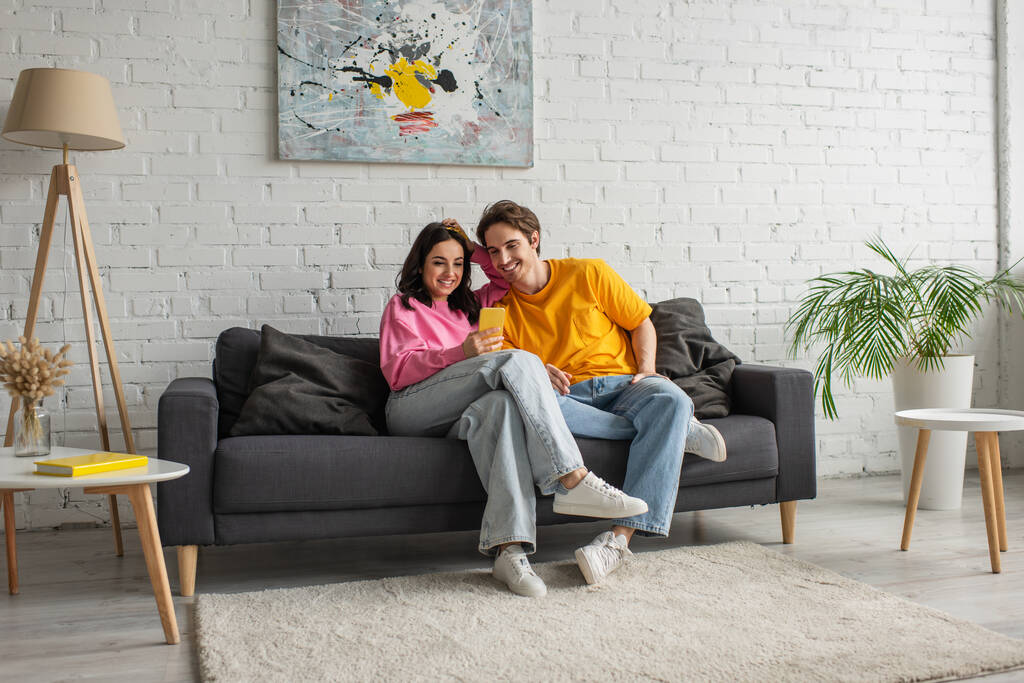улыбающаяся молодая пара сидит на диване и смотрит на сотовый телефон в гостиной - Фото, изображение