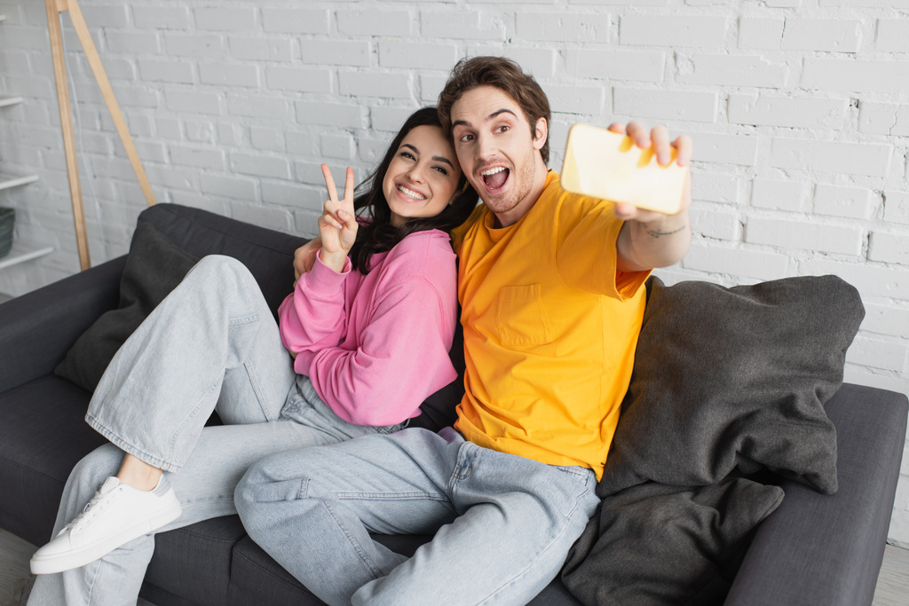 sonriente joven pareja sentada en el sofá y tomando selfie con gesto de paz en la sala de estar - Foto, imagen