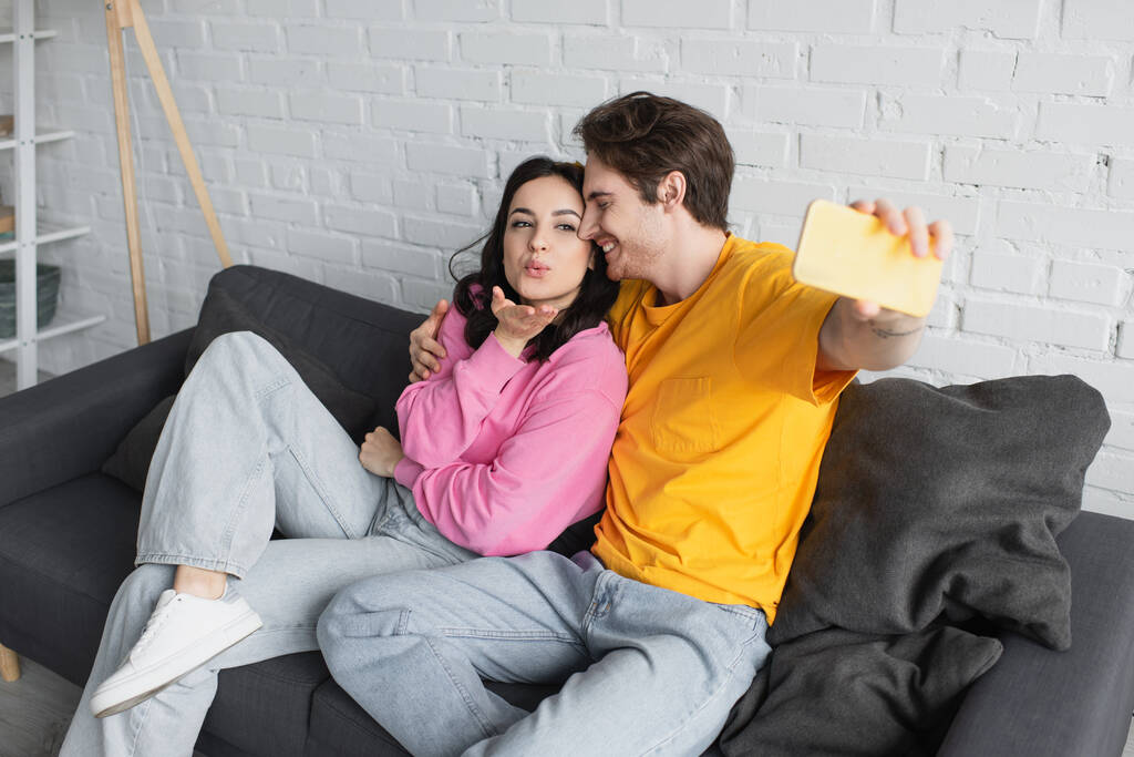uśmiechnięta młoda para siedzi na kanapie i robi selfie z dmuchającym gestem pocałunku w salonie - Zdjęcie, obraz