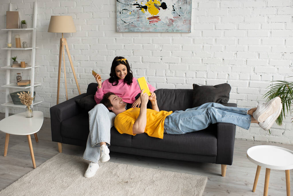 усміхнена молода жінка сидить на дивані з мобільним телефоном біля хлопця, лежить з книгою у вітальні
 - Фото, зображення