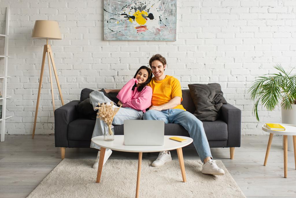 Lächelndes junges Paar sitzt auf Couch und schaut Film auf Laptop im Wohnzimmer - Foto, Bild