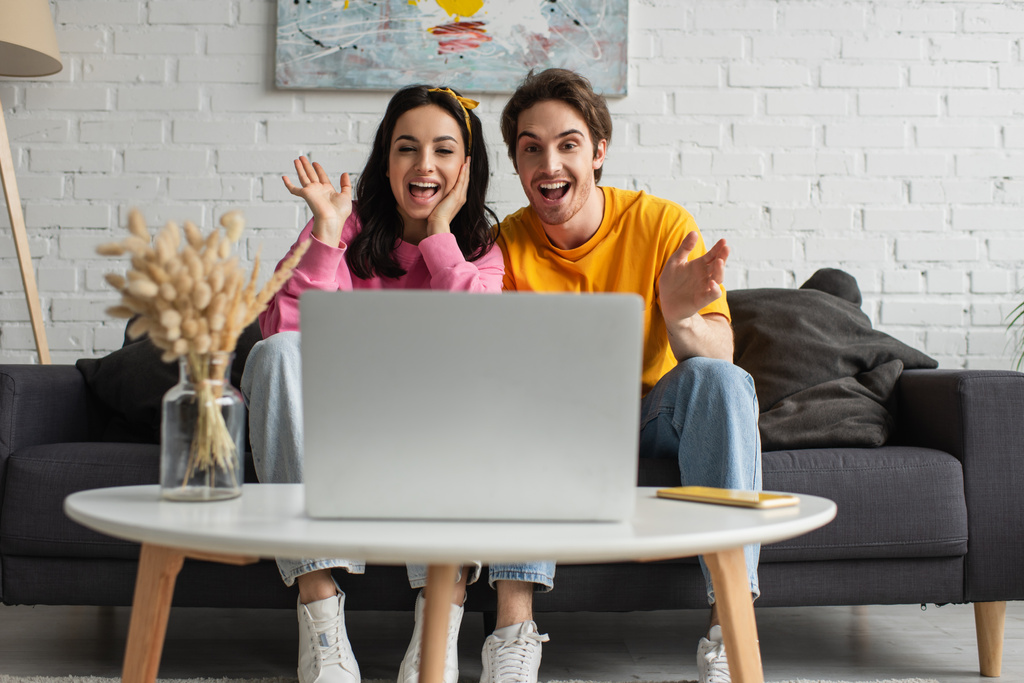 позитивна молода пара сидить на дивані з відкритими ротами і махає руками, маючи відеодзвінок на ноутбук у вітальні
 - Фото, зображення