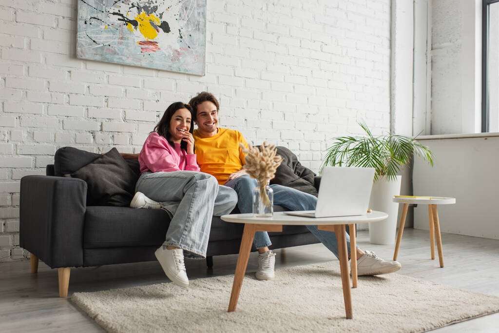 χαμογελαστό νεαρό ζευγάρι κάθεται στον καναπέ και βλέποντας ταινία στο φορητό υπολογιστή στο σαλόνι - Φωτογραφία, εικόνα