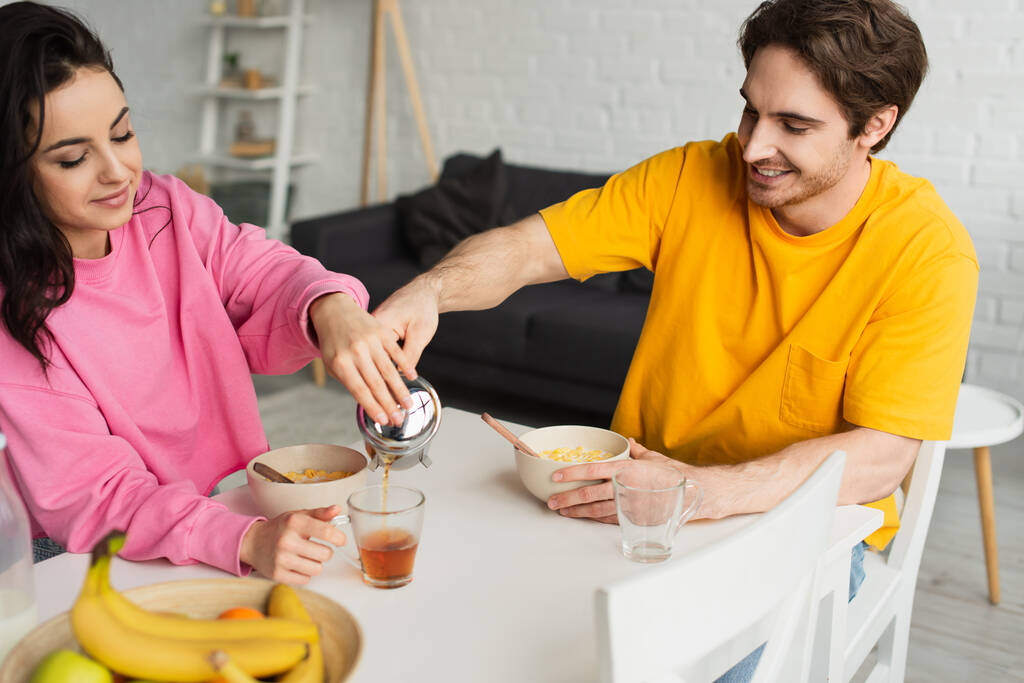 glimlachend jong stel zitten aan tafel met ontbijt en gieten thee van franse pers in beker in woonkamer - Foto, afbeelding