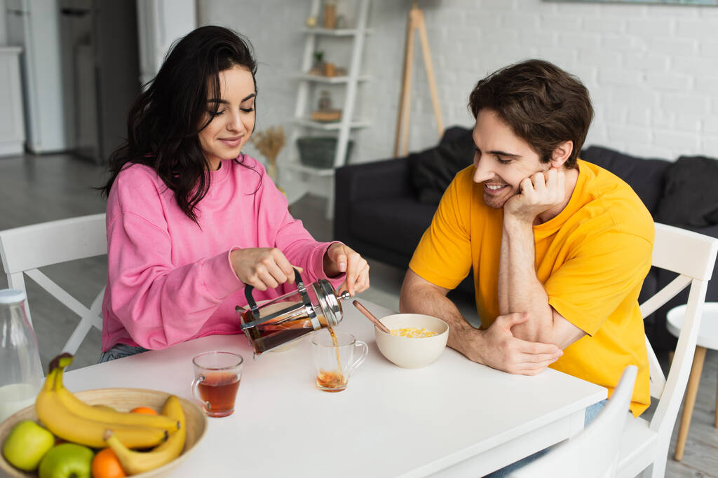 sonriente joven sentada en la mesa cerca del novio con el desayuno y verter té de la prensa francesa en la taza en la sala de estar - Foto, imagen