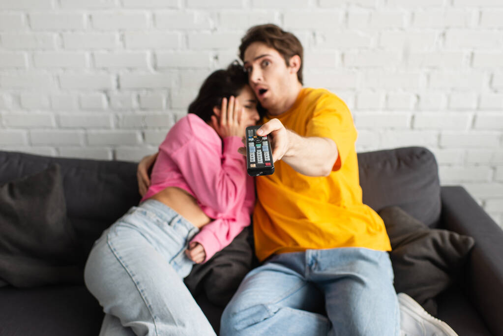 bang jongeman wijzen met afstandsbediening op camera en knuffelen vriendin thuis - Foto, afbeelding