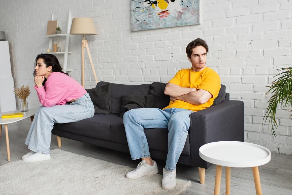 zklamaný mladý muž se zkříženými pažemi sedí na gauči poblíž přítelkyně s rukama blízko obličeje v obývacím pokoji - Fotografie, Obrázek