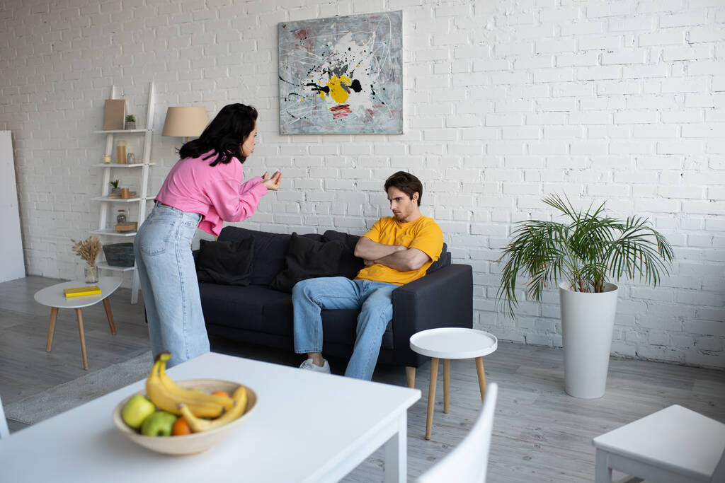 mujer joven enojado gesto con las manos cerca de novio sentado en el sofá con los brazos cruzados en la sala de estar - Foto, Imagen
