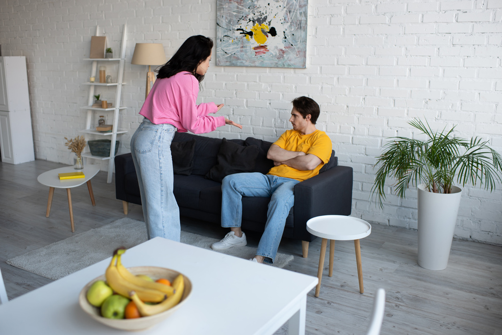 mujer joven enojada gritando con las manos extendidas cerca de novio sentado en el sofá con los brazos cruzados en la sala de estar - Foto, Imagen