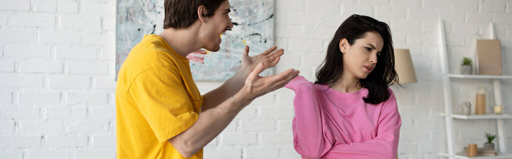 rozzlobený mladý muž křičí s nataženýma rukama blízko přítelkyně v obývacím pokoji, prapor - Fotografie, Obrázek
