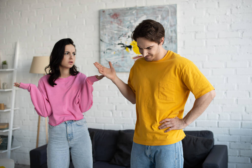 arrabbiato giovane donna in piedi con le mani in aria vicino fidanzato con gesto di rifiuto in soggiorno - Foto, immagini