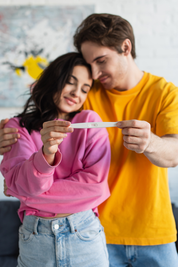 test ciążowy w rękach rozmytej uśmiechniętej młodej pary przytulającej się z zamkniętymi oczami w domu - Zdjęcie, obraz