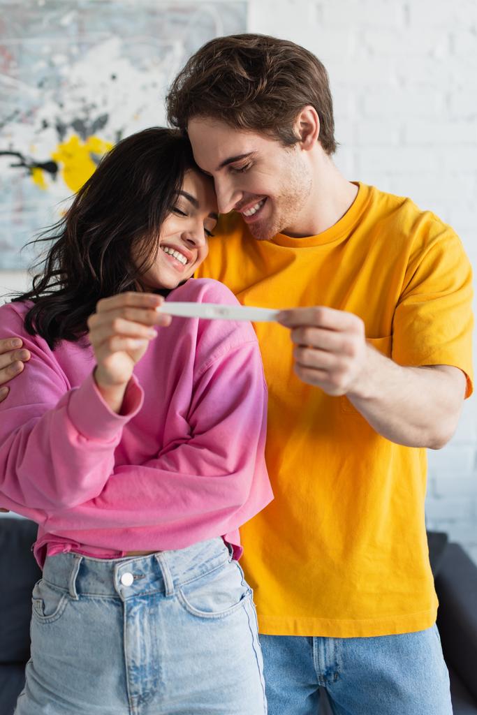 giovane coppia sorridente che si abbraccia a occhi chiusi e tiene il test di gravidanza in mano a casa - Foto, immagini