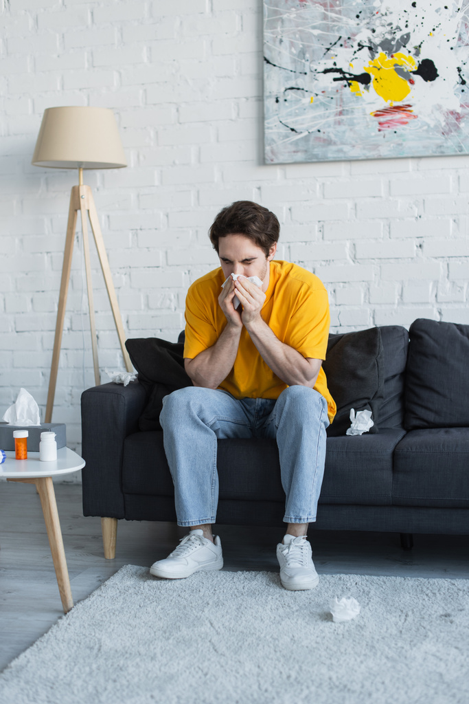 Kranker junger Mann sitzt zu Hause auf Couch und pustet Nase mit Papierserviette - Foto, Bild