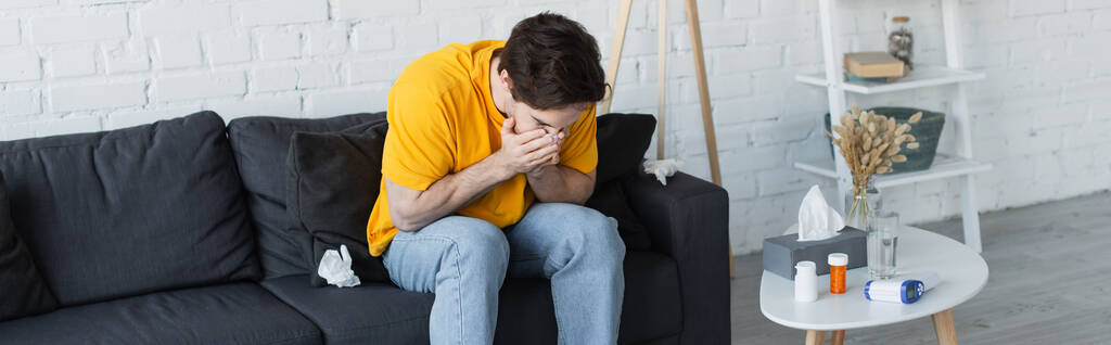 diseased young man sitting on couch and sneezing with hands covering face at home, banner - Photo, Image