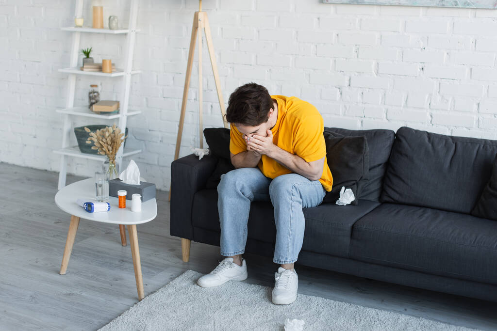 sick young man sitting on couch and sneezing with hands covering face at home - Photo, Image