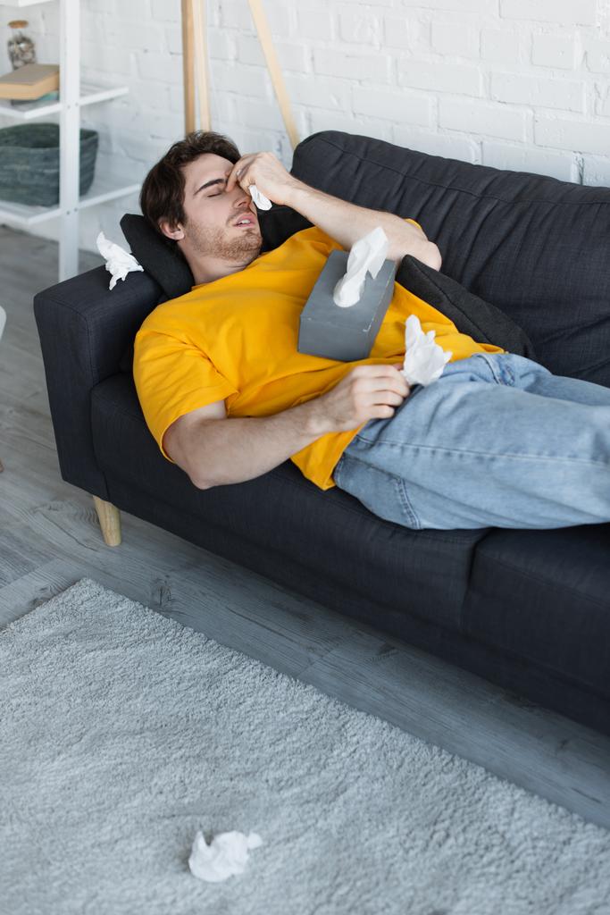 chory młody człowiek leżący na kanapie z ręką w pobliżu głowy i trzymający w domu papierową serwetkę - Zdjęcie, obraz