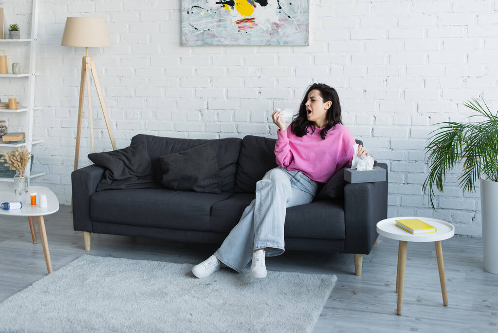 больная молодая женщина сидит на диване и кашляет с бумажной салфеткой в руке дома - Фото, изображение