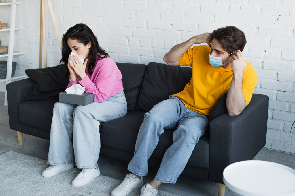 mujer joven enferma sentada en el sofá y sonándose la nariz con servilleta de papel cerca de novio en máscara médica en la sala de estar - Foto, Imagen