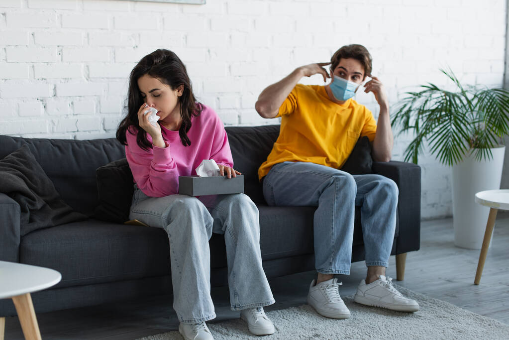 diseased young woman blowing nose with paper napkin near boyfriend sitting on couch and adjusting medical mask at home - Photo, Image