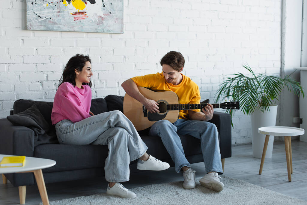 улыбающийся молодой человек, играющий на акустической гитаре рядом с девушкой, сидящей дома на диване - Фото, изображение