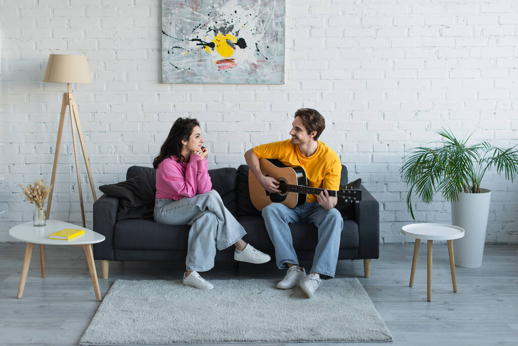 jeune homme souriant assis sur le canapé et jouant de la guitare acoustique près de la petite amie avec les mains près du visage à la maison - Photo, image