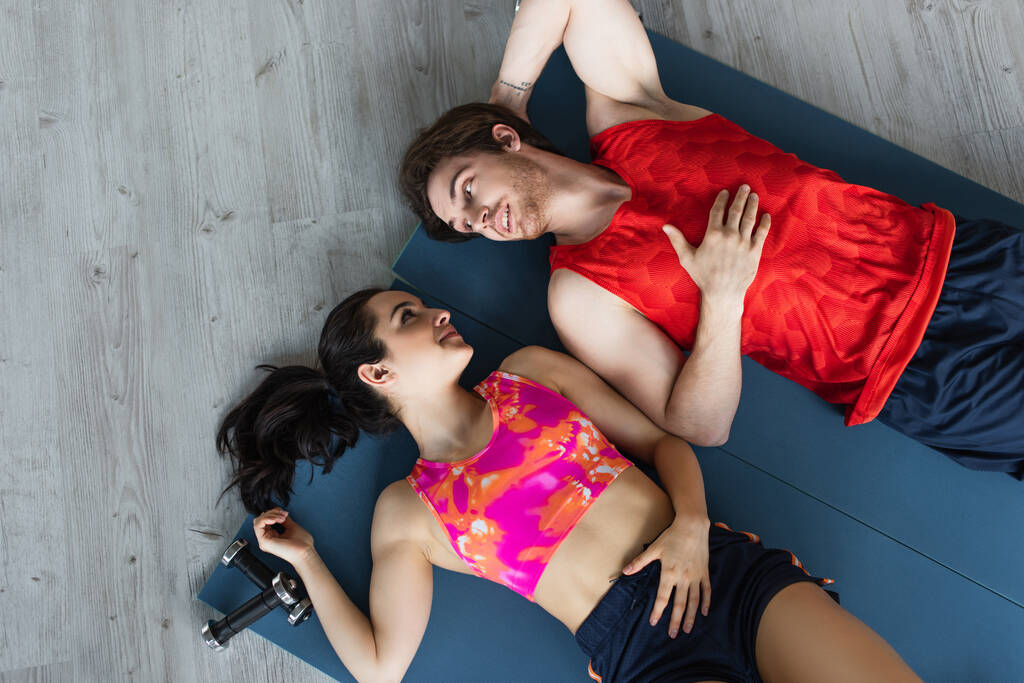 вид на молодую пару в спортивной одежде лежащую на фитнес-ковриках рядом с гантелями и смотрящую друг на друга дома - Фото, изображение