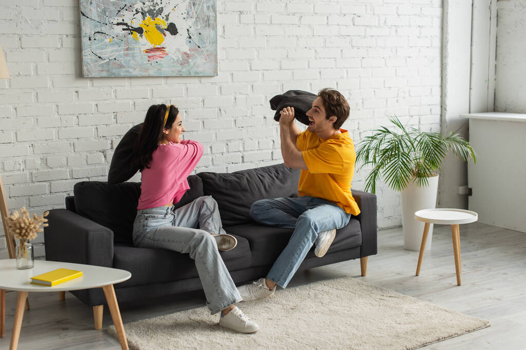 χαμογελαστό νεαρό ζευγάρι που τσακώνεται με μαξιλάρια στον καναπέ στο σαλόνι - Φωτογραφία, εικόνα