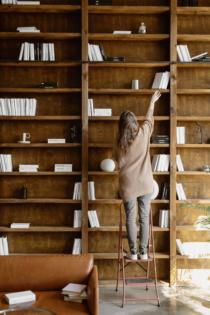 Back view of young woman standing on the step ladder and taking necessary books from the shelf. - Photo, Image