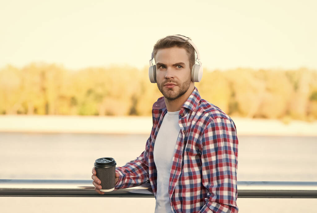 hombre barbudo escuchar música en auriculares digitales y beber café para ir de taza de papel con camisa a cuadros al aire libre, refresco - Foto, imagen
