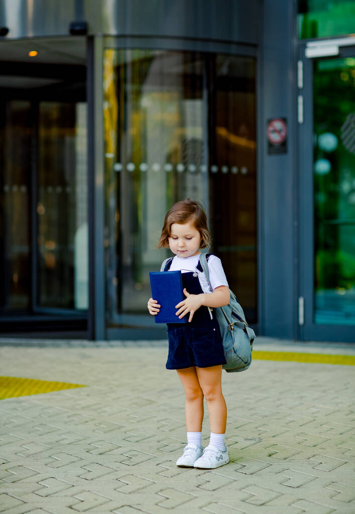 une belle fille caucasienne, une écolière, se tient près de l'école, avec un sac à dos et tenant un journal intime dans ses mains - Photo, image